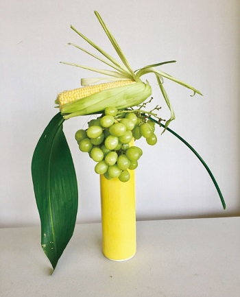 トウモロコシに串を2本挿して、グレープをまたぐように花器に入れる。ハラン（葉）を1枚差し込むと、左右に空間が生まれて奇麗になる